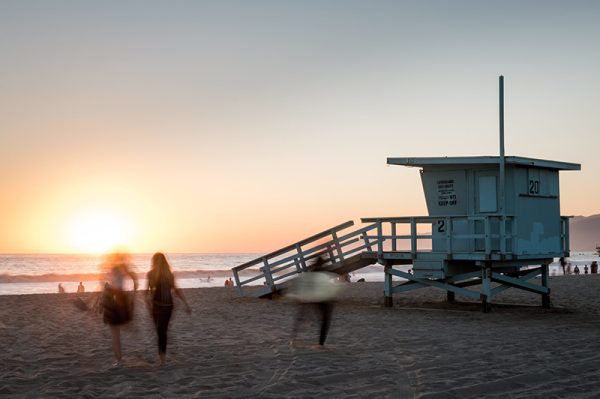People walking on a beach near a lifeguard tower at sunset.