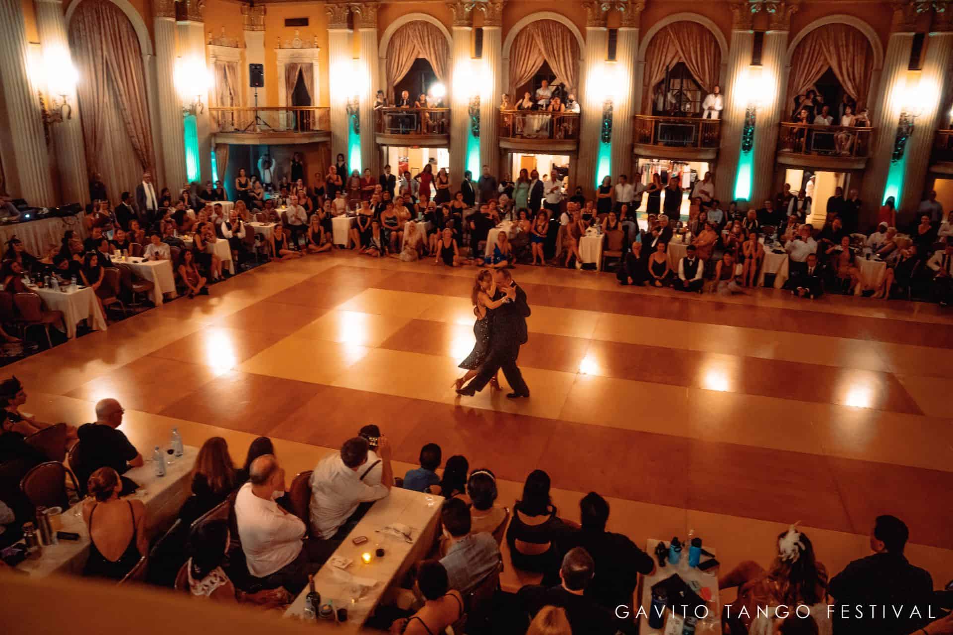 Couple dancing in a ballroom with an audience seated around.