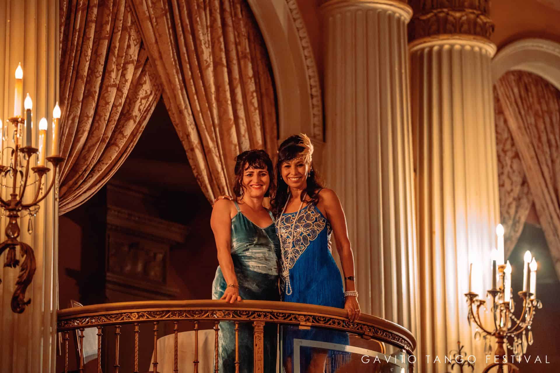 Two women standing on a balcony with warm lighting and elegant curtains.