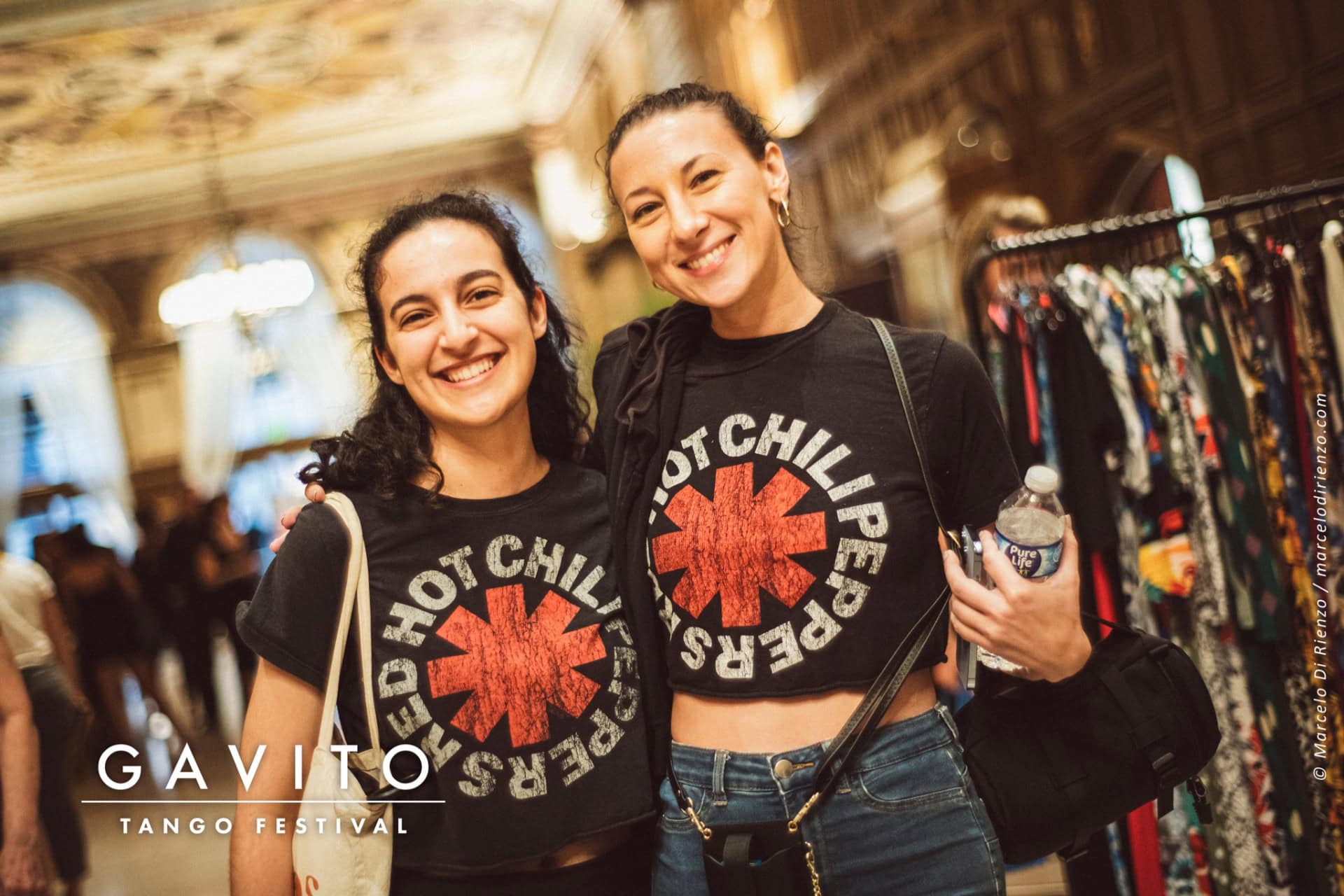 Two women smiling, wearing matching Red Hot Chili Peppers shirts at a lively event.